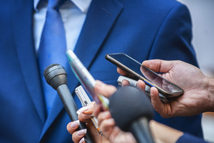 Bild von Mikrofonen, Aufnahmegeräten und Handys, die in Richtung eines Mannes in Anzug gehalten werden.